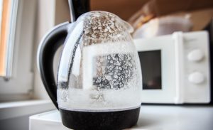 A clear glass electric kettle heavily coated with white limescale mineral deposits, a common effect of using hard water.